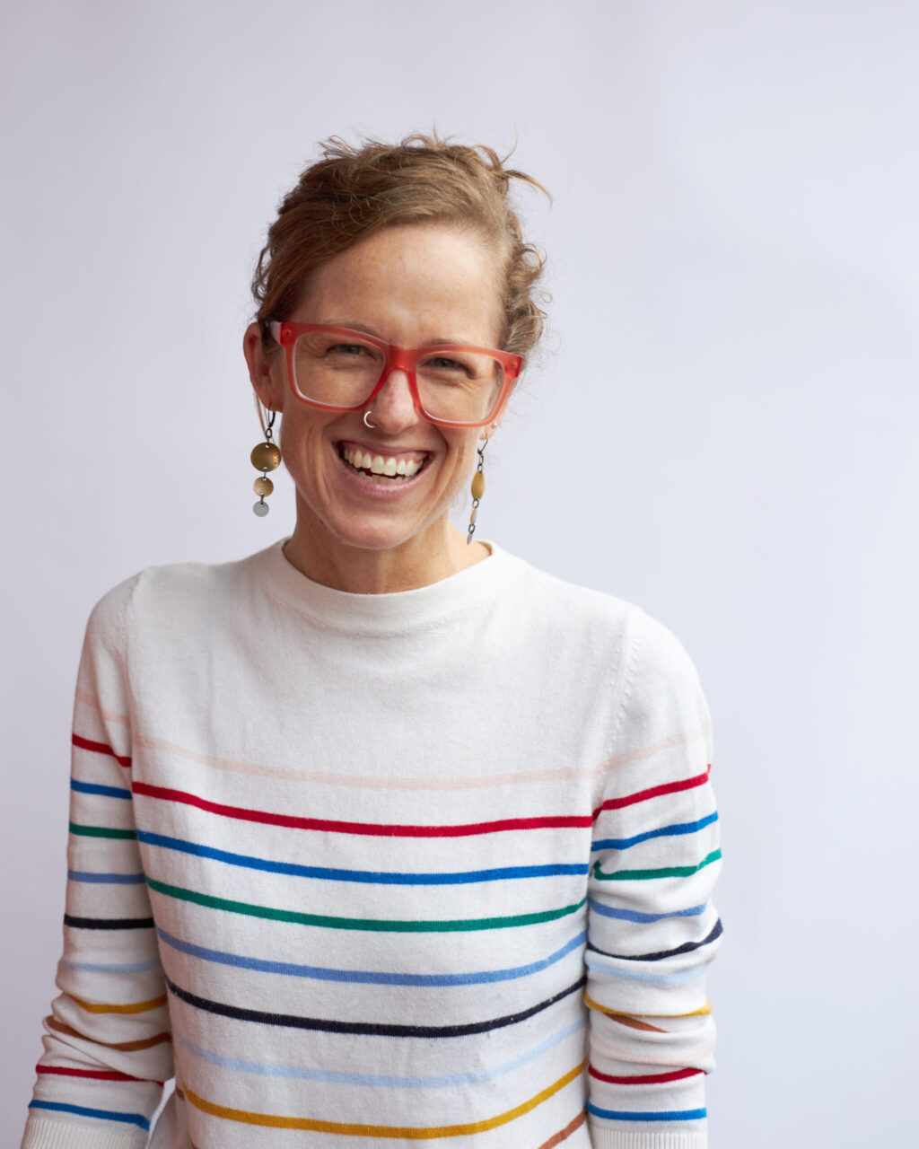 Portrait of Sydney Schaef, Founder of competency-based learning consultancy, CBL Partners, and expert in competency-based education, who is shown smiling and wearing square-framed dark orange glasses and a cream-colored sweater with thin horizontal stripes, each a different color: faded peach, red, blue, green, indigo, black, sky blue, mustard orange, and brown. Background is blurred white.