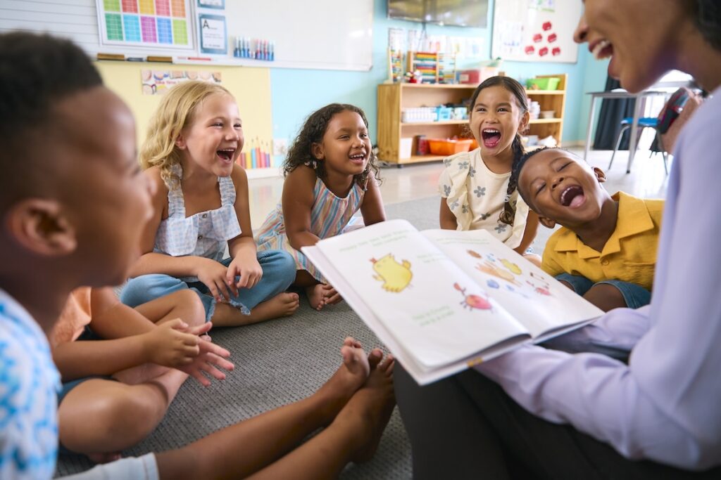 Photograph of a group of students smiling and sitting together in a circle while a teacher, not clearly shown, is reading a book, in a competency-based learning environment.