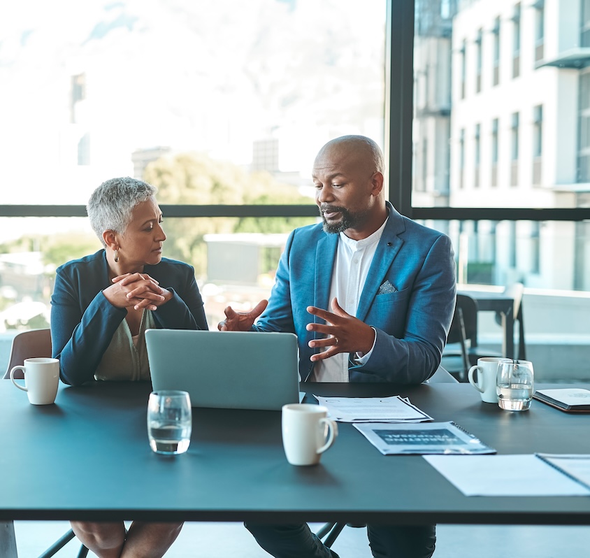 Photograph of two competency-based education leaders dressed in formal clothing are sitting side-by-side at a table with an open laptop, planning together.