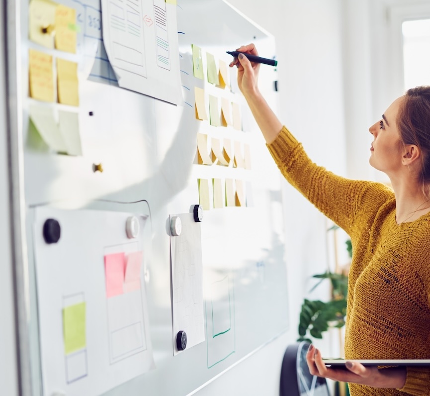 Competency-based learning services in action: A competency-based learning leader is writing on a white board filled with sticky notes and papers held up with magnets. She is wearing a mustard orange long sleeve sweater and holding a laptop in her left hand as she writes.