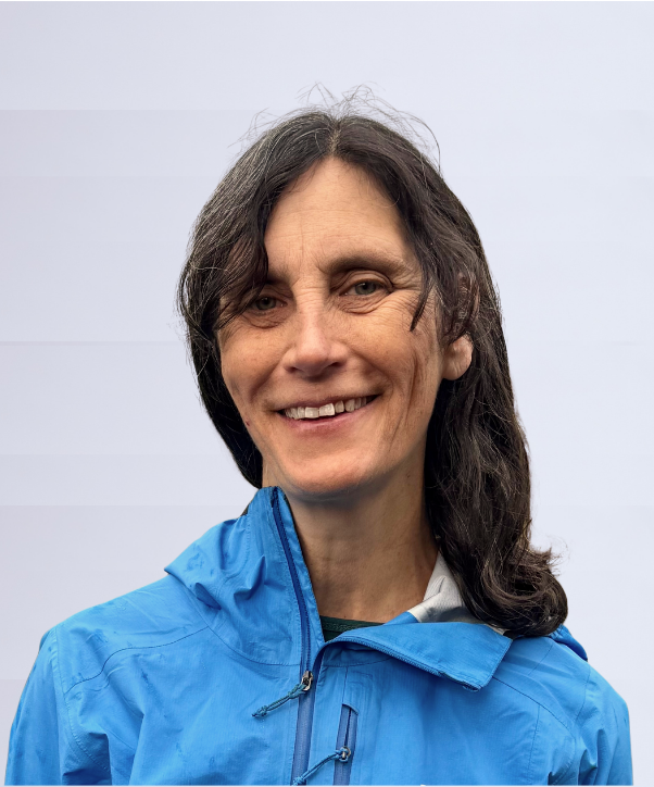 Portrait of Jen Soalt, with a smile, shoulder-length brown hair, a sky blue jacket, and a blurred white background.