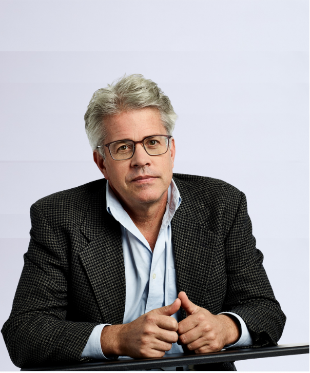A middle-aged man with gray hair and glasses wearing a dark checkered blazer over a light blue shirt, sitting at a table and looking toward the camera with a neutral expression against a plain white background